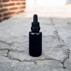 May include: A black glass dropper bottle with a black cap and a long, narrow dropper. The bottle is cylindrical and sits on a gray, textured surface. The background includes a brick wall.