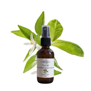 May include: A brown glass spray bottle with a black cap, labelled "Neroli" and "Organic, pure 2 oz." is set against a backdrop of green leaves and white flowers. The bottle contains a clear liquid.