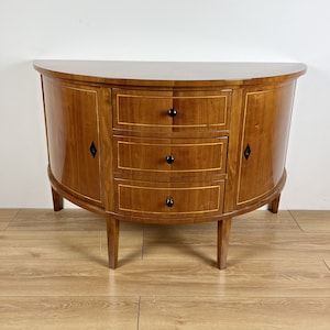 Può includere: Un mobiletto in legno semicircolare con tre cassetti e due ante. Il mobiletto ha una finitura in legno chiaro con accenti in legno scuro. Il mobiletto è posto su un pavimento in legno.