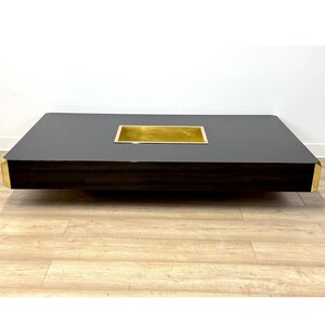 Pode incluir: Uma mesa de centro retangular moderna com um tampo cinza escuro e uma inserção retangular dourada rebaixada. A mesa tem detalhes dourados nos cantos e um perfil baixo.