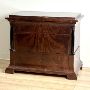 May include: A dark wood chest of drawers with three drawers and black accents. The chest has a curved base and a slightly rounded top.