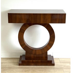Op de afbeelding: Een donkerbruine console in Art Deco-stijl. De tafel heeft een rechthoekig blad met een lade, ondersteund door een groot, cirkelvormig ontwerp met een open centrum. De basis heeft een trapsgewijze, rechthoekige vorm. De houtnerf is zichtbaar.