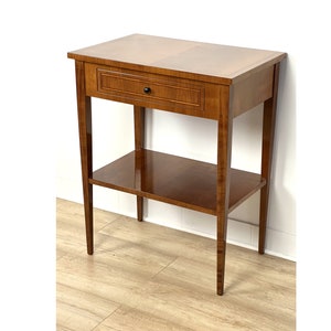 Peut inclure: Une table d'appoint en bois avec un tiroir et une étagère inférieure. La table est de couleur brun clair et est faite de bois massif.