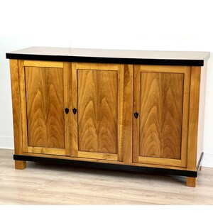 Op de afbeelding: Een houten kast met drie deuren en zwarte afwerking. De deuren zijn gemaakt van licht hout met een houtnerfpatroon. De kast heeft een zwarte bovenkant en staat op een houten vloer.