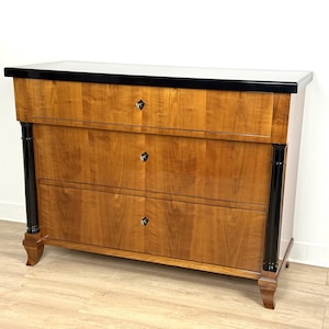 May include: A three-drawer dresser with a black top and black accents on the sides. The dresser is made of wood and has a light brown finish.
