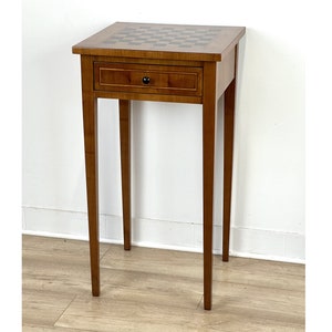 Puede incluir: Una mesa auxiliar de madera con una tapa de tablero de ajedrez y un cajón. La mesa tiene cuatro patas y está hecha de madera marrón claro.