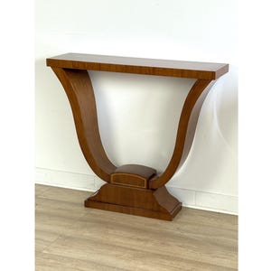 Op de afbeelding: Een houten console tafel met een gebogen voet en een rechthoekig blad. De tafel heeft een warme bruine kleur met een glanzende afwerking. De voet heeft een decoratief design element. Geschikt voor een entree of woonkamer. Ongeveer 120 cm lang.