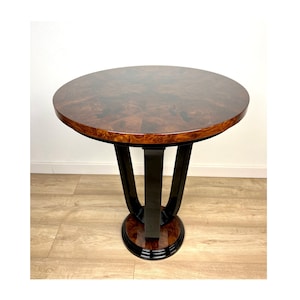 Op de afbeelding: Een ronde, bruine houten tafel met een zwart gelakt onderstel. De tafel heeft een uniek, geometrisch ontwerp en is perfect voor een woonkamer of slaapkamer.