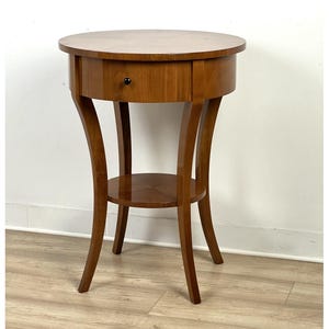 Może przedstawiać: Okrągły, drewniany stolik pomocniczy z szufladą i dolną półką. Stół ma ciepłe brązowe wykończenie i zakrzywione nogi. Szuflada ma małe, ciemne pokrętło. Stół ma około 71 cm wysokości i 61 cm średnicy.
