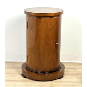 Pode incluir: Um armário cilíndrico de madeira com um tampo claro e uma base escura. O armário tem uma única porta com fechadura e uma pequena maçaneta. O veio da madeira é visível e o acabamento é brilhante.