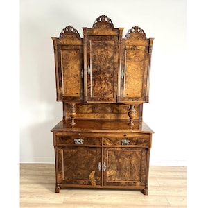 May include: A large, antique wooden cabinet with a dark, patterned finish. The cabinet features multiple doors and drawers, ornate carvings, and decorative hardware. The upper section has three doors, and the lower section has two doors and two drawers.
