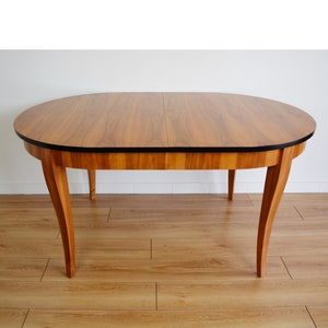Puede incluir: Una mesa de comedor ovalada de madera con un borde negro y cuatro patas. La mesa está hecha de madera clara y tiene un diseño sencillo.