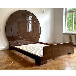 Op de afbeelding: Een donkerbruin bed in Art Deco-stijl met een groot, cirkelvormig hoofdbord. Het hoofdbord heeft concentrische cirkels en een glanzende afwerking. Het rechthoekige bedframe heeft een lattenbodem. Het bed staat tegen een witte muur.