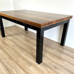 Puede incluir: Una mesa de comedor rectangular de madera con una base pintada de negro y una tapa de madera marrón. La mesa tiene una hoja que se puede extender para hacerla más larga.