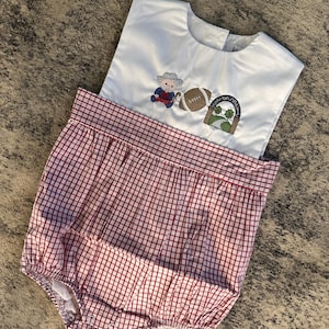 Puede incluir: Un body blanco para bebé con la parte inferior a cuadros rojos y blancos. La parte superior tiene un diseño bordado de un balón de fútbol, un duende y una figura sentada. El body está diseñado para un bebé o un niño pequeño.