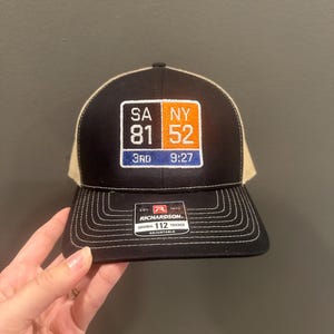 Könnte beinhalten: Eine schwarze und beige Trucker-Cap mit einem rechteckigen gestickten Aufnäher mit dem Text SA 81, NY 52, 3RD und 9:27. Das Vorderteil ist schwarz mit weißen Nähten am Schirm und einem schwarzen Aufkleber mit der Aufschrift Richardson 112 Trucker.
