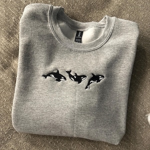 May include: A light grey crewneck sweatshirt featuring an embroidered design of three black and white orca whales swimming in a horizontal line across the centre of the chest. The garment has a ribbed collar and a soft fleece interior lining.