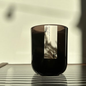 May include: A dark brown glass candle jar with a white label that reads "fumo" and "10 oz | New York, New York". The candle is sitting on a light-colored, slatted surface. The background is a white wall with shadows.