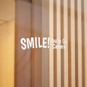 Pode incluir: Um autocolante de vinil branco aplicado numa superfície de vidro com o texto SMILE! You're On Camera numa fonte negrito sem serifas. A palavra SMILE! é apresentada num tamanho maior, seguida pelo texto mais pequeno You're On Camera.
