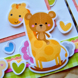 Op de afbeelding: Een gele giraffe met bruine vlekken en een bruine teddybeer die op haar rug rijdt. De giraffe houdt een roze bloem in haar mond. De achtergrond is een geruit patroon.