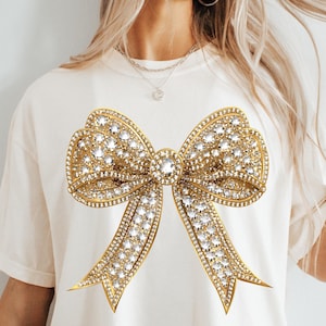 May include: A cream-colored short-sleeve t-shirt featuring a large, central graphic of a gold-toned bow. The bow design is embellished with numerous clear, sparkling rhinestones that create a textured, metallic appearance across the fabric.