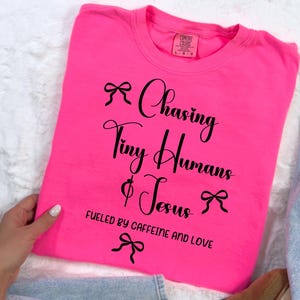 Può includere: Una maglietta rosa acceso con la scritta "Chasing Tiny Humans & Jesus" in corsivo nero, accentuata da grafiche a fiocco. La frase "Fueled by Caffeine and Love" è stampata sotto. La maglietta è piegata su una superficie bianca.