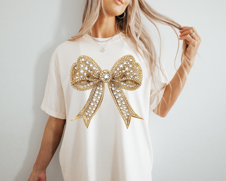 May include: A cream-colored short-sleeve t-shirt featuring a large, central graphic of a gold-toned bow. The bow design is embellished with numerous clear, sparkling rhinestones that create a textured, metallic appearance across the fabric.