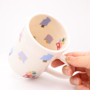 Könnte beinhalten: Weißer Keramikbecher mit Henkel, verziert mit bunten Baum- und Blumenmustern. Auch das Innere des Bechers weist das gleiche Design auf. Der Becher wird schräg gehalten, wodurch das Design zur Geltung kommt.