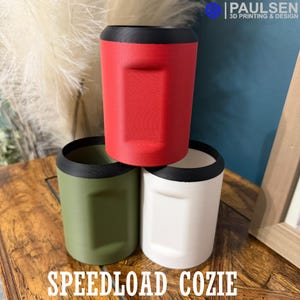 Puede incluir: Tres portavasos impresos en 3D en rojo, verde y blanco, cada uno con un borde negro. El portavasos rojo está apilado encima de los portavasos verde y blanco. El texto "SPEEDLOAD COZIE" está en la parte inferior de la imagen.