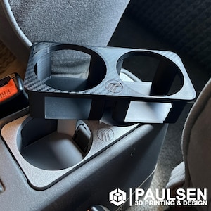 May include: A black 3D printed cup holder with a carbon fiber pattern. The cup holder is designed to fit into a car's center console. The cup holder has two slots for cups.