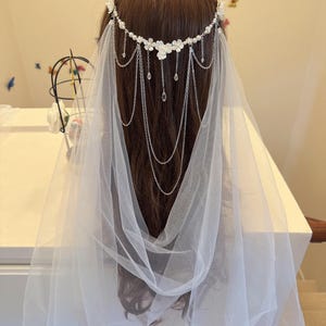 K&ouml;nnte beinhalten: Ein wei&szlig;er Braut Schleier mit einem zarten Blumen- und Ketten-Haarschmuck. Der Haarschmuck ist mit kleinen wei&szlig;en Bl&uuml;ten und Kristall-Akzenten verziert.