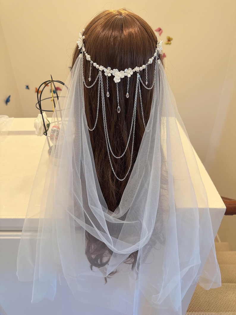 K&ouml;nnte beinhalten: Ein wei&szlig;er Braut Schleier mit einem zarten Blumen- und Ketten-Haarschmuck. Der Haarschmuck ist mit kleinen wei&szlig;en Bl&uuml;ten und Kristall-Akzenten verziert.