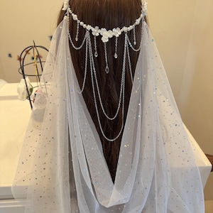 K&ouml;nnte beinhalten: Wei&szlig;er Braut Schleier mit einem zarten Blumen- und Ketten-Haarschmuck. Der Schleier ist aus einem durchsichtigen Stoff mit einem dezenten Glitzern.