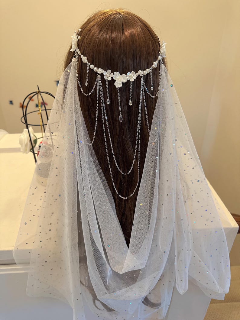K&ouml;nnte beinhalten: Wei&szlig;er Braut Schleier mit einem zarten Blumen- und Ketten-Haarschmuck. Der Schleier ist aus einem durchsichtigen Stoff mit einem dezenten Glitzern.
