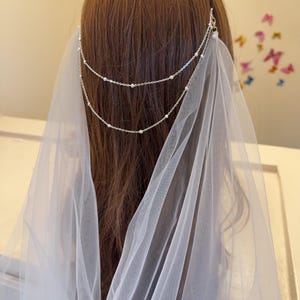 May include: A white bridal veil with a silver chain headpiece featuring small beads. The veil is made of soft, sheer fabric and drapes elegantly over the shoulders.