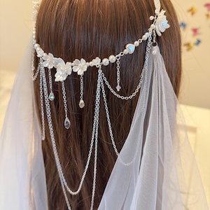 K&ouml;nnte beinhalten: Ein wei&szlig;er Blumenhaarschmuck mit einer zarten Kette und Perlenakzenten. Der Haarschmuck ist mit kleinen wei&szlig;en Bl&uuml;ten und einer einzigen gr&ouml;&szlig;eren Bl&uuml;te verziert. Die Kette ist aus Silber und hat einen zarten, femininen Look. Der Haarschmuck ist perfekt f&uuml;r eine Hochzeit oder einen anderen besonderen Anlass.
