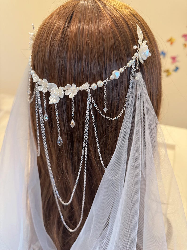 K&ouml;nnte beinhalten: Ein wei&szlig;er Blumenhaarschmuck mit einer zarten Kette und Perlenakzenten. Der Haarschmuck ist mit kleinen wei&szlig;en Bl&uuml;ten und einer einzigen gr&ouml;&szlig;eren Bl&uuml;te verziert. Die Kette ist aus Silber und hat einen zarten, femininen Look. Der Haarschmuck ist perfekt f&uuml;r eine Hochzeit oder einen anderen besonderen Anlass.