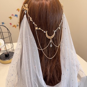 以下が含まれることがあります： 三日月型のチャームとクリスタルビーズが付いたゴールドのヘアチェーンが、グリッターのアクセントが付いた白いチュールベールから垂れ下がっています。