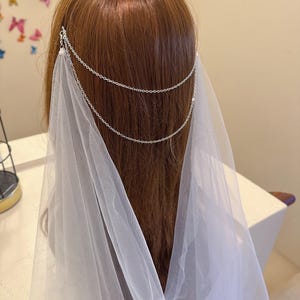 Op de afbeelding: Een witte bruids sluier met een zilveren ketting hoofdband. De sluier is gemaakt van zacht tule en heeft een delicaat, vloeiende ontwerp. De ketting hoofdband is versierd met kleine, glinsterende kralen.