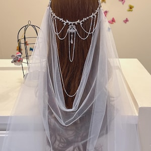 May include: A white tulle veil attached to a silver-toned metal headpiece featuring a delicate leaf motif, intricate filigree center, and multiple draped chains. A single clear teardrop-shaped crystal hangs from the center of the metal ornament.
