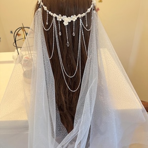 K&ouml;nnte beinhalten: Ein wei&szlig;er Braut Schleier mit einem zarten Blumen- und Ketten-Haarschmuck. Der Schleier ist aus einem durchsichtigen, gepunkteten T&uuml;llstoff gefertigt.