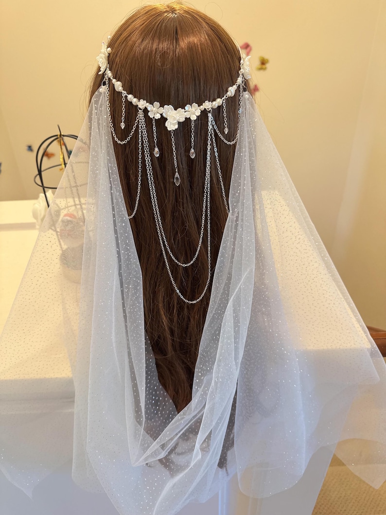 K&ouml;nnte beinhalten: Ein wei&szlig;er Braut Schleier mit einem zarten Blumen- und Ketten-Haarschmuck. Der Schleier ist aus einem durchsichtigen, gepunkteten T&uuml;llstoff gefertigt.
