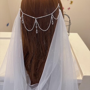 May include: A silver-toned headpiece with dangling crystal accents is draped over a head of brown hair. A sheer white veil with sparkling details flows down the back, creating a dreamy, ethereal look. The headpiece is a delicate accessory.