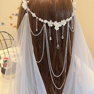 K&ouml;nnte beinhalten: Ein wei&szlig;er Braut-Haarschmuck mit Perlen- und Blumenakzenten und einer langen Kette aus silbernen Perlen. Der Haarschmuck wird mit einem wei&szlig;en Schleier getragen.
