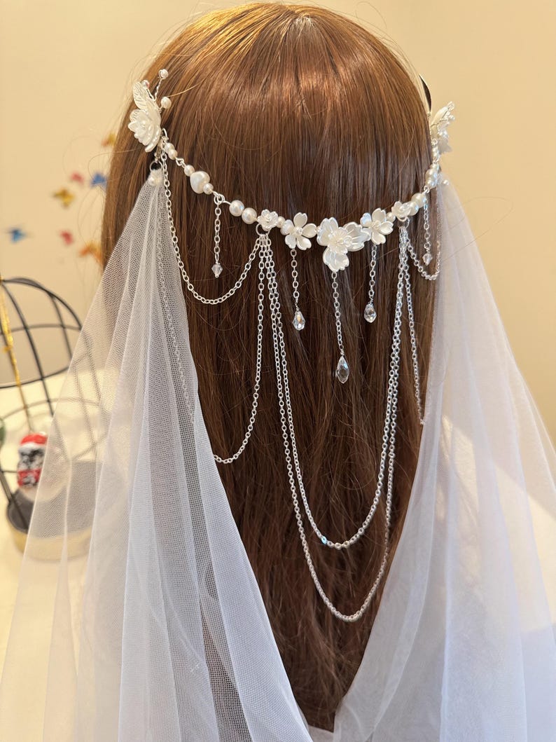 K&ouml;nnte beinhalten: Ein wei&szlig;er Braut-Haarschmuck mit Perlen- und Blumenakzenten und einer langen Kette aus silbernen Perlen. Der Haarschmuck wird mit einem wei&szlig;en Schleier getragen.