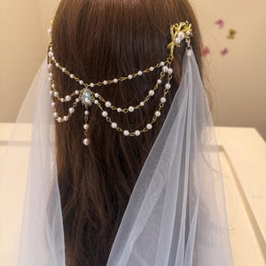 以下が含まれることがあります： 真珠とクリスタルをあしらったゴールドのブライダルヘアアクセサリー。白いチュールベールに付いています。