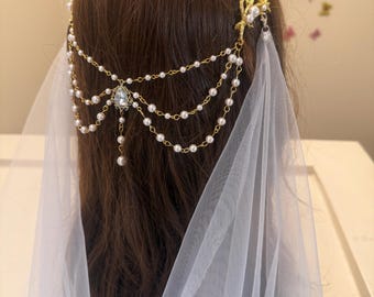 Puede incluir: Un tocado de novia dorado con un diseño de perlas y cristales, unido a un velo de tul blanco.