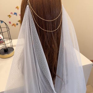 May include: A bridal headpiece featuring a gold chain and a sheer white veil. The veil is dotted with small, sparkling accents. The headpiece is designed to be worn at the back of the head, with the veil cascading down.