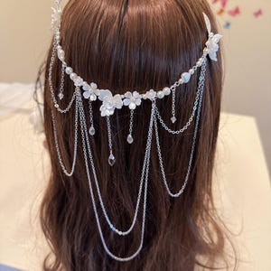 K&ouml;nnte beinhalten: Ein wei&szlig;er Blumenhaarkamm mit einer silbernen Kette und Perlenakzenten. Der Kamm wird im braunen Haar einer Frau getragen.