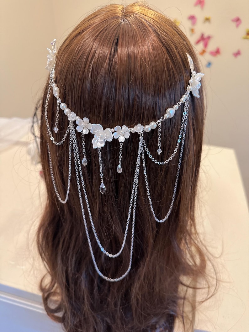 K&ouml;nnte beinhalten: Ein wei&szlig;er Blumenhaarkamm mit einer silbernen Kette und Perlenakzenten. Der Kamm wird im braunen Haar einer Frau getragen.